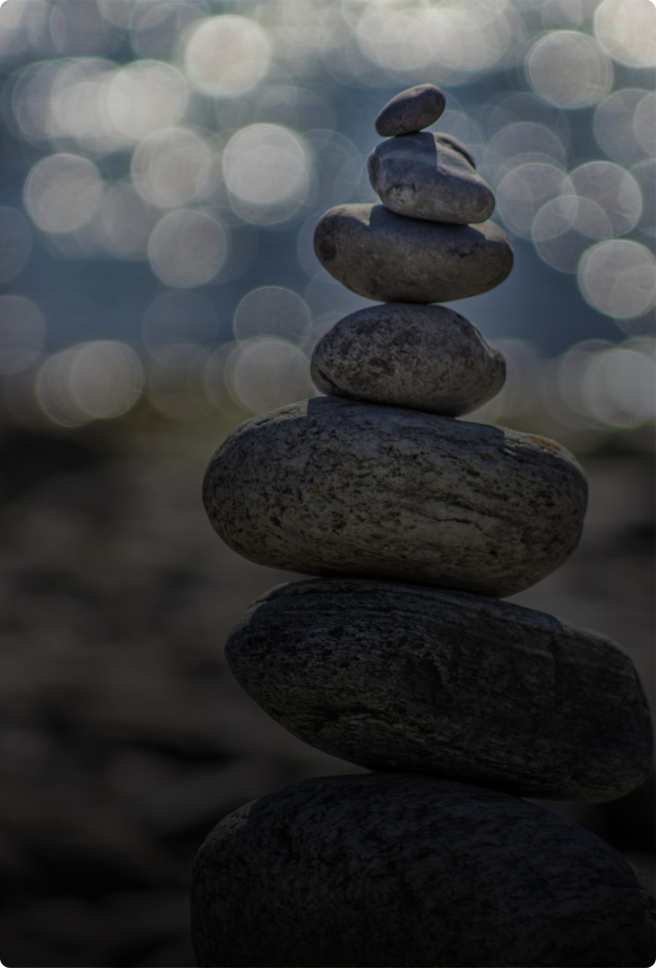 A cairn of seven smooth grey stones stacked in decreasing size on a dark beach, with deep slate-blue water and soft circular light reflections glowing behind it.